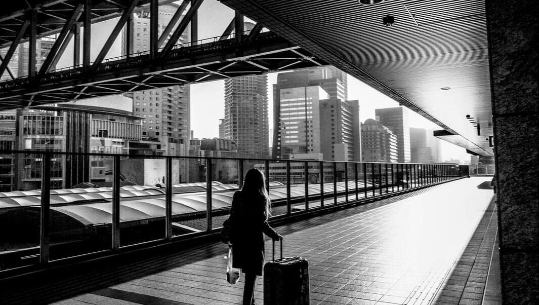 Black and white image of a long-haired person with a luggage at a long, empty platform with a cityscape in the background.  Photo by masahiro miyagi on Unsplash  